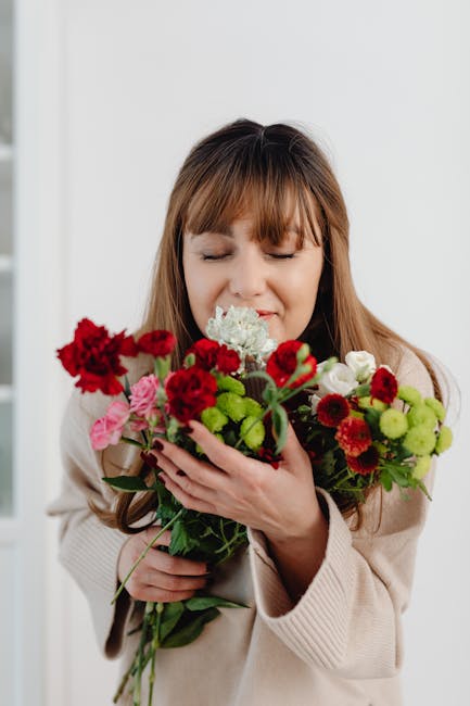 A woman with long light brown hair and a gentle expression is holding a vibrant bouquet of fresh flowers, including red, pink, white, and green blooms, with lush green foliage. The bouquet features va