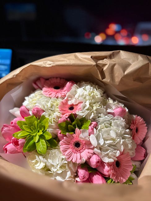 A bouquet of fresh white hydrangeas and pink gerbera daisies, with a few green succulent leaves, wrapped in brown craft paper, held in a person's hand against a dark, blurred background with streetlig