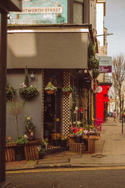 A cozy floral shop on a street corner features an outdoor display of vibrant fresh flowers arranged in wooden crates and pots, including bright pink, purple, and yellow blooms. The entrance is flanked