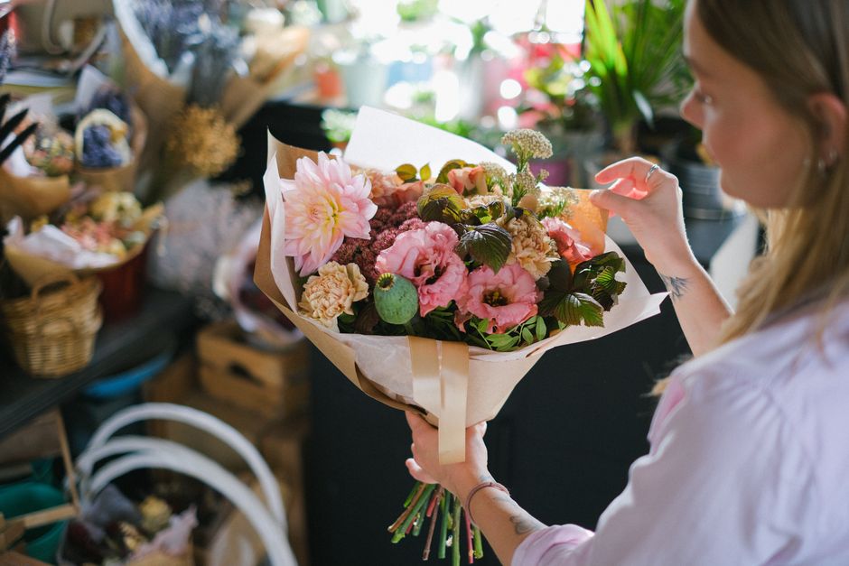 A woman with blonde hair, wearing a light pink top, holding a large bouquet of fresh flowers from flowerdeliverybexleyheath.co.uk. The bouquet includes pink dahlias, light pink lisianthus, pale peach 
