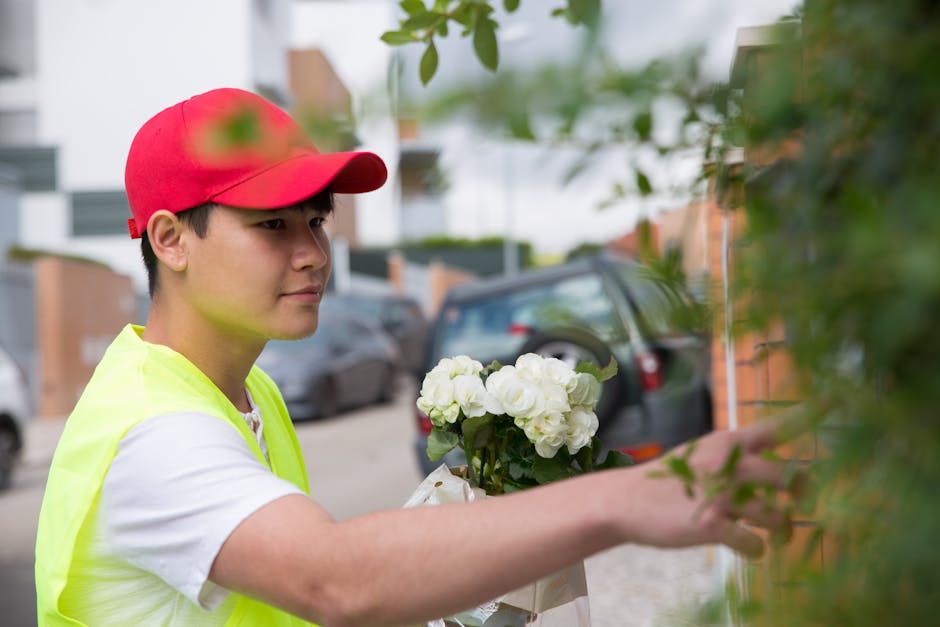 A young male delivery person wearing a red cap and a neon yellow vest is placing a bouquet of white hydrangeas wrapped in white paper against a brick wall, with a background of parked cars and residen