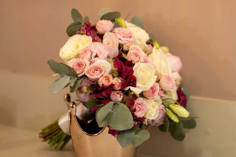 A floral bouquet arrangement featuring pale pink and white roses, complemented by deep burgundy flowers and green eucalyptus leaves, displayed in a shiny silver vase. The bouquet is placed on a light-