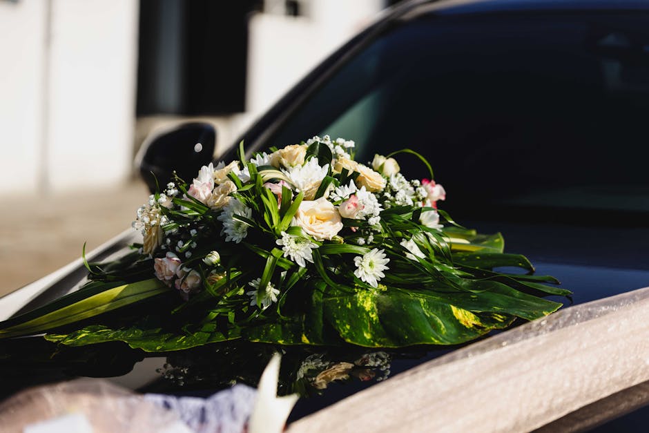 A delicate floral arrangement placed on the hood of a black car, featuring a bouquet of fresh white roses, pale pink lisianthus, small white daisies, and sprigs of baby's breath, all surrounded by lus