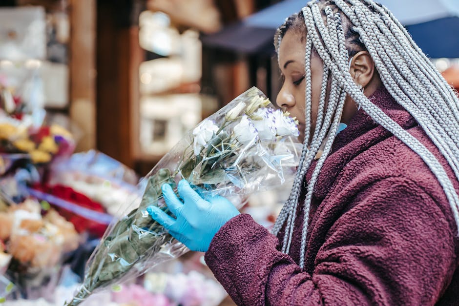 A woman with silver braided hair, wearing a maroon fleece jacket and blue gloves, is holding a floral bouquet wrapped in transparent plastic. The bouquet contains predominantly white flowers with some