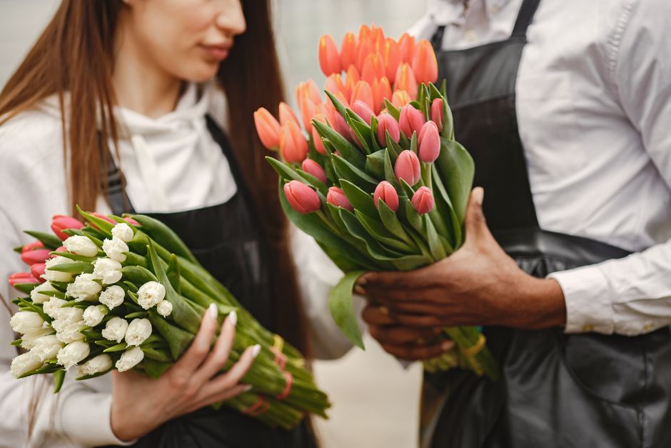 A florist from flowerdeliverybexleyheath.co.uk is handing over two bouquets of fresh tulips, one with pink tulips and the other with white tulips, both with lush green leaves. The customer, a woman wi