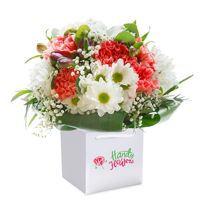 Standing regally in a spotless white square gift bag, this striking bouquet showcases the signature style of Bexleyheath's local flowershops. At its heart bloom plush, ruffled carnations in coral, peach, and bright red-textures as rich as a velvet ribbon, colours as lively as any spring morning along the paths of Danson Park. Peppered throughout, radiant white daisy chrysanthemums with glowing lime-green centers infuse the display with a crisp, uplifting spirit. Threaded lightly among the main flowers, delicate baby's breath brings a sense of softness, much like the gentle clouds that often drift over Bexleyheath's rooftops. Glossy, deep green foliage forms a lush surrounding collar, anchoring the arrangement in natural elegance. A closer look reveals a few closed purple-tipped buds, ready to unfurl new life into the bouquet. Each element is precisely placed to create a harmonious, full silhouette-one that feels ready-made for celebration or sharing across Bexleyheath's friendly neighbourhoods. The sleek, paper gift bag, finished with strong white ribbon handles, is as practical as it is stylish, ideal for hassle-free delivery. With professional photographic clarity, the image captures every aspect of freshness and quality. This bouquet's artistry and vibrant tones make it a perfect floral gift for birthdays, milestones, or simply sending heartfelt warmth throughout Bexleyheath.