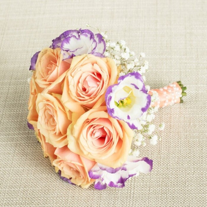 Inviting touch and admiration, this hand-crafted bouquet rests elegantly on a soft linen canvas, its subtle warmth echoing Bexleyheath's welcoming vibe. The roses are a symphony in peach and soft apricot, with petals unfurling to reveal creamy, delicate blushing hearts-a favourite palette for local summer weddings. Countering this tender warmth, crisp white lisianthus blossoms punctuate the bouquet, their charming ruffled petals brushed in bright violet-each edge looking expertly painted for an artisanal touch, much like the bespoke details found in Bexleyheath's town centre shops. Baby's breath clusters are scattered throughout, their feathery lightness lending a whimsical air, evocative of celebrations on a gentle breeze at Danson Park. Each element is arranged with intentional balance; the stems nestle together, carefully wrapped in a smooth ribbon of peachy-pink, its surface accented by subtle pearl beads, echoing the refined details beloved by Bexleyheath brides. Soft daylight warms the scene, making this arrangement ideal for a heartfelt occasion at a community venue or a significant family gathering. Every nuance embodies the careful craftsmanship and warmth that have made Bexleyheath florists a trusted choice for those seeking beauty with a personal, local touch.
