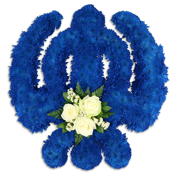 An impressive Sikh Khanda floral tribute fills the frame, its bold religious symbolism translated into a rich tapestry of royal blue petals. Set against a fresh white background, the entire Khanda emblem has been shaped with precision: the central, upright sword; the perfect circular chakkar surrounding it; and the two symmetrical kirpans sweeping gracefully outward. All are built from a multitude of small, densely placed chrysanthemum-like blooms, each floret contributing to a continuous, velvety surface. The intense blue colour exudes solemnity and honour, echoing the reverent atmosphere often felt at Sikh funeral services and remembrance gatherings in Bexleyheath. At the base of the central circular area, a delicate accent of white and green provides a softer focal point. Three open white roses, with layered petals that curl gently at the edges, rest among sprays of tiny white filler flowers such as gypsophila, all supported by glossy, dark green foliage. This contrasting posy doesn't overwhelm the Khanda but instead enhances it, adding a sense of peace and calm to the powerful blue design. Fine shadows along the outer edges of the emblem suggest a substantial, almost sculptural presence, revealing the careful handwork involved in constructing the tribute. Likely prepared by an experienced local florist familiar with Sikh customs, this floral Khanda would be suitably placed at a gurdwara, beside a coffin, or at a memorial venue in Bexleyheath. It conveys respect not only for the faith but also for the individual being remembered, combining visual impact with deep cultural significance.