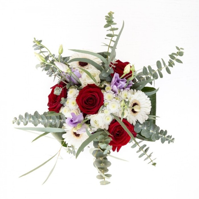 As a Bexleyheath florist, I see this bouquet as a tender collaboration of nature's finest forms and hues-confident, yet understated in its seasonal appeal. The bouquet's vivid red roses evoke enduring love and steadfast devotion, their spiraled forms catching the soft light that so often fills homes near Danson Lane. Around them, creamy white gerbera daisies with bold, dark centers offer a classical counterpoint, their linear petals framing the roses and drawing the gaze outward. Silvery arcs of eucalyptus rise above, their delicate leaves casting moving shadows and introducing a breezy, aromatic freshness. Scattered among the showcase blooms, blush-lilac lisianthus impart a contemporary whisper-a soft echo of garden evenings in Bexleyheath. Puffs of ivory filler flowers and clusters of small chrysanthemums add tactile interest and fullness, their textures inviting gentle exploration. The foundation of wide, emerald green leaves encircling the clear glass vase emphasizes both structure and poise; beneath, the stems rest in pure water, a testament to craftsmanship and freshness. This bouquet is at home in celebrations-be they anniversaries, new beginnings, or gentle remembrance-gracing tables along Broadway or sent as a caring gesture to your loved ones in Bexleyheath.