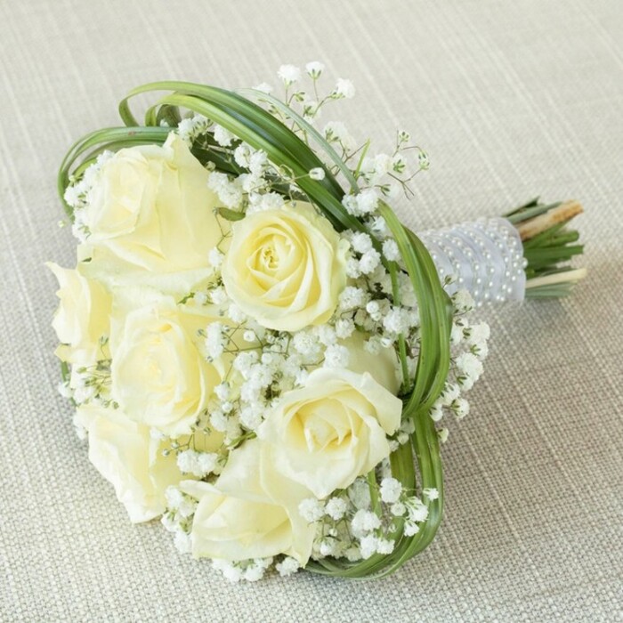 Framed against a neutral beige fabric-softly textured, like those seen at Bexleyheath's chic event spaces-this bridal bouquet exudes a harmonious blend of classic romance and modern artistry. The heart of the bouquet features an array of luscious creamy white roses, each meticulously selected at the ideal stage-from youthful, tightly curled buds to fully blossomed flowers revealing layers of opulent petals. Generous tufts of delicate white baby's breath (Gypsophila) intersperse the roses, lending airy volume and a lace-like texture that evokes the gentle atmospheres of Bexleyheath's historic townscapes. Slender strands of vivid green bear grass loop artistically around the bouquet, providing lively contrast and a contemporary frame for the blooms. The stems are expertly gathered and wrapped in lustrous, light silver ribbon, finished with stately rows of smooth, radiant pearls that both secure and embellish the arrangement. The cut stem ends spill just beneath this refined wrap, a nod to the fresh, locally sourced blooms. Lovingly arranged by Flower Delivery Bexleyheath, this bouquet serves as a perfect symbol for meaningful milestones-whether exchanging vows by the gardens of Bexleyheath Village, or toasting an anniversary in a sunlit home-bringing together skilled floristry, timeless purity, and a taste of the local community.