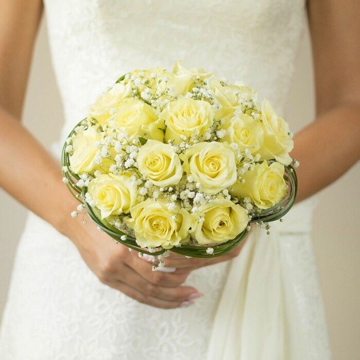 A close-up, taken from chest level in a light-filled Bexleyheath venue, frames a bouquet that encapsulates classic romance and refined craftsmanship. Tightly packed yellow roses, their petals luminous and perfectly sculpted, form a sumptuous dome, each bloom seeming to glow with quiet joy. Dotting the arrangement, petite white sprays of baby's breath offer a whimsical touch, their delicate shapes and airy brightness enhancing the vibrant roses. At the bouquet's base, dark green, possibly wire-wrapped or leaf-formed bands swirl around, anchoring the bouquet and reflecting the gentle beauty of Bexleyheath's garden squares. Resting just above the intricate folds of a pristine white gown, the hands holding the bouquet are elegant and steady, with neat, pale nails and the sparkle of a wedding ring catching the light. The softly blurred background hints at lively celebration, reminding one of Bexleyheath's bustling wedding venues. This bouquet not only celebrates love-it embodies the indispensable artistry and sense of occasion found in Bexleyheath's local floral traditions.