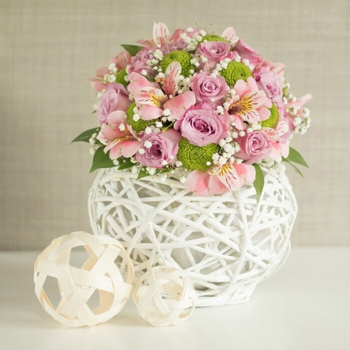Bringing together the sophistication and local heritage of Bexleyheath, this radiant floral display exudes elegance and gentle charm. The generously rounded bouquet is a harmony of pastel pink and lavender roses, their textured petals showcased in full bloom and accompanied by light pink alstroemeria bearing their distinctive, softly patterned markings. Bright lime green button chrysanthemums punctuate the arrangement, injecting contemporary energy that lifts the gently blended palette, while clusters of baby's breath lend an airy, ethereal glow. All these exquisite florals nestle within a white, open-weave vase whose interlaced strips and bulbous silhouette reference the modern, artisanal influences that punctuate Bexleyheath's creative spirit. Nearby, two decorative spheres-each woven from pale, natural material with textures both airy and robust-are thoughtfully placed on the seamless white surface, mirroring the community's affection for hand-crafted design. The arrangement sits before a light, subtly textured backdrop that sets off the joyous blooms, creating a sense of welcoming celebration perfect for wedding tables or spring gatherings. Whether marking a milestone, sending a heartfelt gift, or simply brightening a Bexleyheath home, this floral center display embodies freshness, balance, and the artistry for which Flower Delivery Bexleyheath is known.