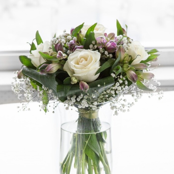 In this image, a refined bouquet of creamy white roses, lavender-hued alstroemeria, and delicate baby's breath is arranged in a clear, cylindrical glass vase, glowing softly in diffused natural light that could easily be streaming through a Bexleyheath window on a mild afternoon. The roses, fully open yet flawless, sit at the centre of the design, their petals unfurling in layered swirls that create a plush, romantic heart to the bouquet. Around them, stems of alstroemeria in gentle shades of lilac, lavender, and muted purple emerge, some blossoms fully open and others still in elongated buds, edged with green sepals that hint at fresh growth. Fine sprays of white gypsophila are tucked between the larger blooms, their tiny star-like flowers catching the light and giving the arrangement a soft, cloud-like halo. Deep green foliage encircles the flowers, with glossy, broad leaves intertwined with more slender, arching stems, adding texture, contrast, and a feeling of natural abundance. Through the transparent glass, a neat bundle of green stems is visible, fanning out in clear water that underscores the bouquet's freshness and the care taken in conditioning each stem. The composition is rounded and balanced, with no harsh angles, as though expertly crafted by a local florist in Bexleyheath who understands how to blend traditional romance with contemporary simplicity. The mood is one of quiet elegance and gentle reassurance, making this arrangement suitable for sending sympathy, celebrating a milestone, or brightening a home near Danson Park or along the tree-lined roads that run off Bexleyheath Broadway. It feels like a bouquet chosen to speak softly yet sincerely, letting the flowers carry sentiments of love, remembrance, or heartfelt congratulations.