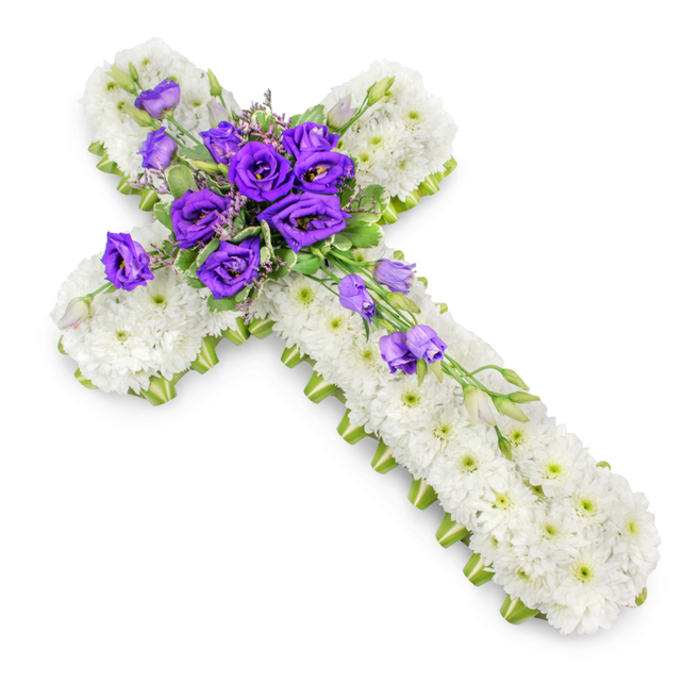 This detailed funeral cross arrangement is captured against a pure white background, allowing every element of the design to stand out with clarity. The cross itself is constructed from a tightly packed blanket of white spray chrysanthemums, each flower small and pristine, their petals overlapping to create a soft, velvety field of white. Subtle green eyes at the centre of each bloom add fine detail and prevent the design from feeling flat. Along the outer edges, a narrow lime-green ribbon traces the entire outline, hugging the arms and base of the cross. This ribbon border provides a crisp frame, emphasising the strong lines of the cross and lending a contemporary touch to a traditional tribute. At the centre, a rich cluster of deep purple lisianthus forms the focal point. The lisianthus flowers, some fully open and others just beginning to unfurl, show layered, ruffled petals that add depth and a sense of luxury. Threaded between them are sprigs of light green foliage and small filler flowers, which introduce a natural, airy feel and draw the eye gently outwards into the white chrysanthemum base. From this central spray, a trailing line of lisianthus stems extends down the right side of the cross, dotted with additional purple blooms and buds to create a graceful, diagonal cascade. Taken from a slightly elevated angle, the photograph highlights texture, shape and the delicate interplay of colours, while soft lighting casts a faint shadow beneath the cross to give it dimension. The overall impression is one of calm devotion and quiet beauty, a style of floral cross that families in Bexleyheath might choose for a church altar or crematorium chapel to honour a loved one with faith, love and enduring respect.