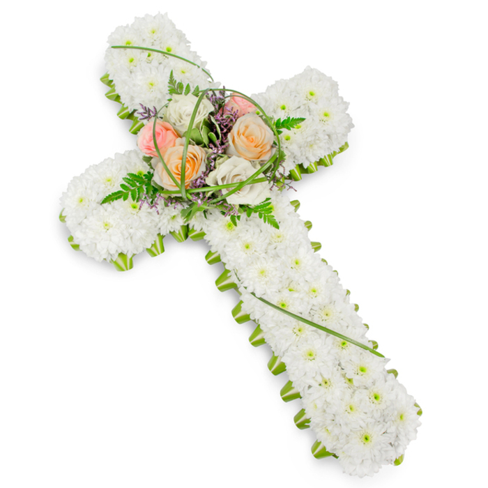 This image shows a refined floral cross, a classic funeral tribute set against a stark white background that highlights every detail of the design, much like a well-lit chapel space in Bexleyheath. The cross itself is three-dimensional and thoroughly carpeted with tiny white cushion chrysanthemums, or comparable small blooms, arranged so closely that no gaps are visible. The result is a soft, plush surface of pure white petals that conveys serenity, purity, and a sense of gentle rest. Encircling the entire outline is a delicate ribbon of bright green foliage, sharply defining the arms and base of the cross and offering a fresh, living contrast that enhances the shape's clarity. At the central junction of the cross, a raised cluster of larger blooms forms a prominent focal point. Several roses in harmonising tones of soft peach, warm apricot, blush pink, and creamy ivory unfold in layered spirals, their subtle hues bringing warmth and emotional depth to the arrangement. These roses are interwoven with feathery fern fronds, fine strands of vivid green grass, and tiny purple filler blossoms, which add nuanced colour accents and a light, airy texture. The overall lighting is clean and diffuse, revealing the structure and balance of the cross without distraction. This floral tribute reflects the kind of thoughtful, hand-crafted sympathy arrangement often created by experienced florists for families in Bexleyheath, suitable for display in local churches, funeral homes, or at gravesides around the area. It serves as a symbolic expression of Christian faith, remembrance, and enduring affection for a loved one who has passed.
