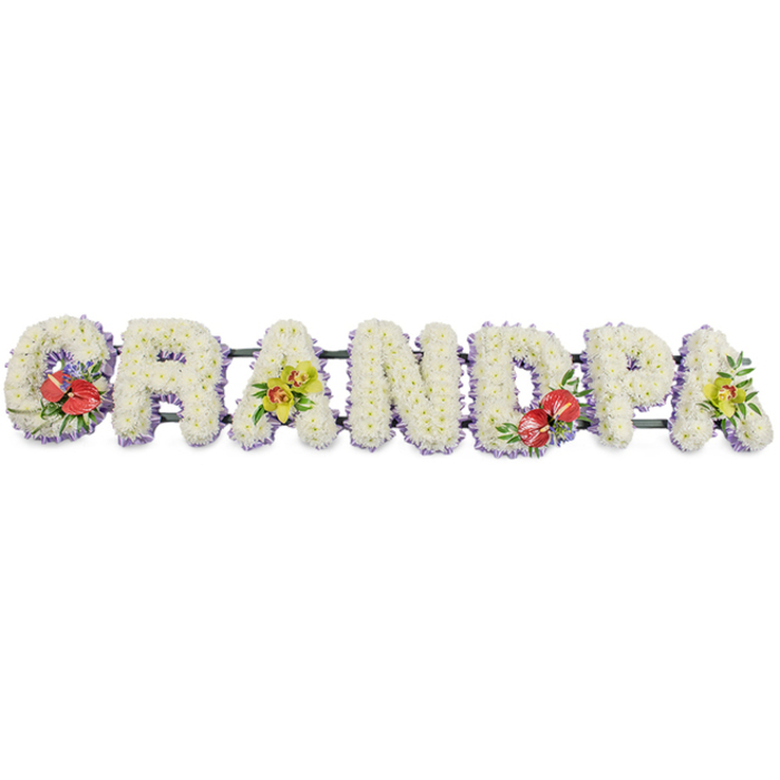 A meaningful funeral flower arrangement spells the word "GRANDPA" in bold block capitals, stretching horizontally across the frame against a simple, uncluttered white background. Each letter looks solid and reassuring, formed from dense clusters of small white chrysanthemums that completely cloak the underlying frame. The chrysanthemums are arranged with great care so their petals overlap into a soft, pillowy texture, conveying a sense of comfort and serenity, like a white quilt of blossoms. The even colouring of creamy white blooms gives the tribute a peaceful, unified appearance. Around the base of every letter runs a band of soft lavender ribbon, providing a cool, pastel border that gently accentuates the shapes without overpowering the design. The letters are connected on a dark green framework that stays largely tucked away behind the flowers, giving stability while allowing the word to be read clearly from a distance-ideal for placing at the front of a Bexleyheath church or crematorium chapel. Four key letters feature artfully arranged accent flowers: the "G" and "D" display sprays of deep red anthuriums, their waxy, heart-shaped spathes and central spadices framed by lush green foliage and hints of filler blooms, adding depth and intensity. The first and last "A"s balance those deeper tones with sprays of yellow-green orchids, their long, graceful petals and buds bringing a fresh, modern touch. The tribute feels personal and heartfelt, the kind of bespoke piece families in Bexleyheath might order from Flower Delivery Bexleyheath to honour a grandfather whose presence anchored family gatherings, shopping trips along the Broadway, or quiet afternoons in local gardens. It stands as a floral expression of gratitude, respect, and enduring love.