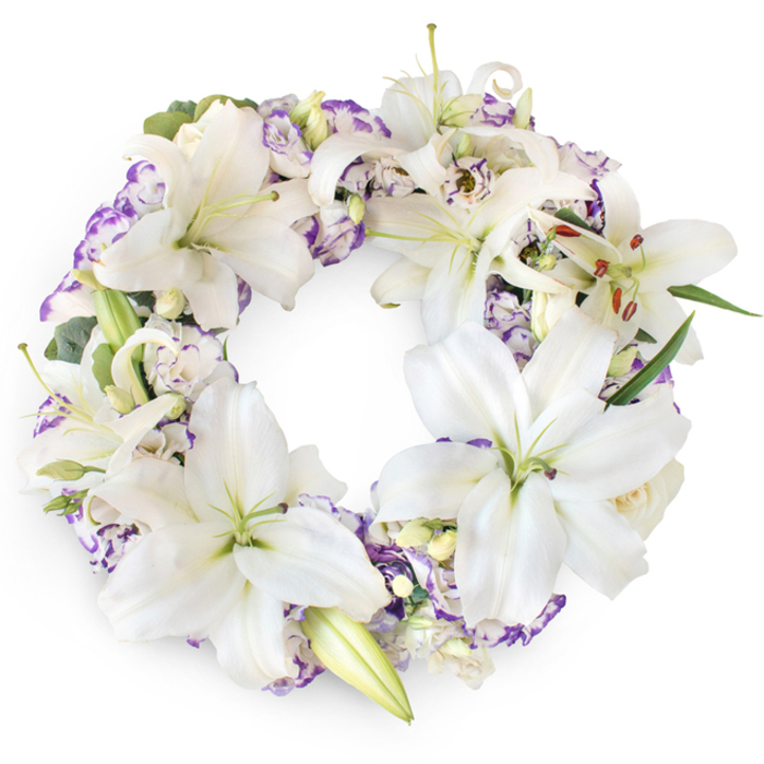 A meticulously arranged funeral wreath forms a complete, unbroken ring of white and lavender flowers, carefully set against a clean white backdrop that highlights its purity and detail. The focal blooms are striking white Oriental lilies, their petals broad and luminous, fanning out in gentle arcs. Some lilies open fully, revealing pale green throats and a central cluster of stamens tipped with cinnamon-brown anthers, while others are still enveloped in elegant, elongated buds, adding a sense of natural progression to the design. Between these prominent flowers, clusters of Lisianthus appear, their petals softly layered and ruffled, each one traced with subtle lilac and purple edging that looks almost hand-painted. These purple accents are thoughtfully spaced around the wreath, creating rhythmic pockets of colour that draw the eye along the circular form. Small creamy buds, reminiscent of newly formed roses, are tucked here and there, adding youthful touches among the more expansive blooms. Supporting everything is a discreet base of mixed greenery, including deep green leaves and eucalyptus-style sprigs whose muted silver-green tone adds depth and coolness without distracting from the lilies and Lisianthus. The visual impression is of a calm, dignified tribute-an embodiment of sympathy, respect, and enduring love. Within Bexleyheath, this wreath would be at home gracing a funeral service near Danson Park or accompanying a memorial in a chapel off Broadway, its quiet elegance reflecting the community's care for the bereaved. The thoughtful craftsmanship suggests the steady hand of a local Bexleyheath florist who understands how important a single floral tribute can be in moments of farewell.