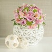 Bexleyheath's spirit of community and celebration shines through this vibrant, hand-assembled floral centerpiece, styled atop a crisp white surface against a soft, textured beige background. The centerpiece overflows with pastel-hued roses-gentle pinks and soothing lavenders exuding timeless romance-intertwined with delicate pink alstroemeria, their slender petals streaked and tipped with elegant markings reminiscent of a Bexleyheath garden in full bloom. Lively lime green button chrysanthemums peek playfully from the bouquet, their compact texture providing a sparkling highlight, while clouds of white baby's breath contribute an ethereal finishing touch, floating gently between the larger blooms to create softness and balance. The unique white vase, made from slender, intertwining strips in a latticework fashion, offers both rustic warmth and modern sophistication-a nod to the creative traditions cherished in local shops. Beside the vibrant arrangement, two pale, woven decorative spheres, one larger and one smaller with open, airy patterns, reinforce the sense of thoughtful craftsmanship and connection to the community's artistic flair. With its harmonious blend of colour, texture, and local character, this piece is perfectly suited as a wedding table centerpiece or a heartfelt gift to bring joy and tranquillity to a Bexleyheath home, lovingly delivered to your door.